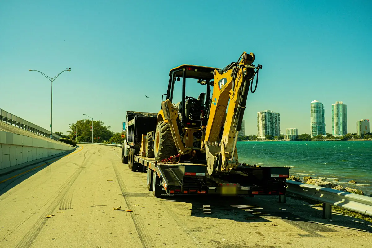 A yellow backhoe loader chained onto a black equipment trailer behind a dump truck, stopped on a causeway road beside turquoise water with high-rise towers in the distance.
