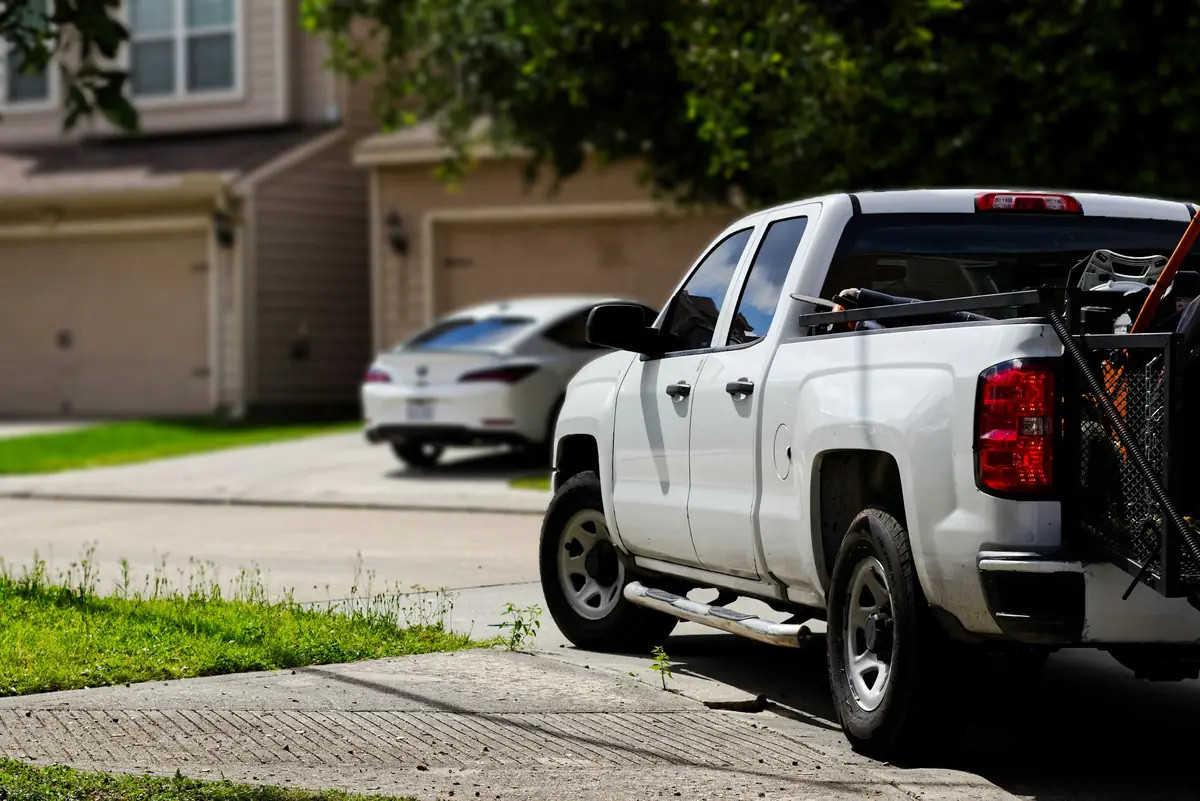 A white pickup truck with lawn-care tools on a rack in its bed, parked at the curb of a suburban street with houses and a white car behind.